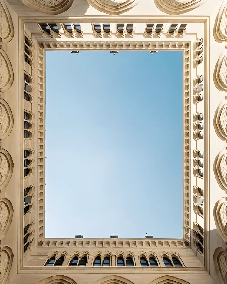 Pátio interno de um edifício histórico em pedra clara, visto de baixo, com janelas em arco emoldurando um recorte de céu azul